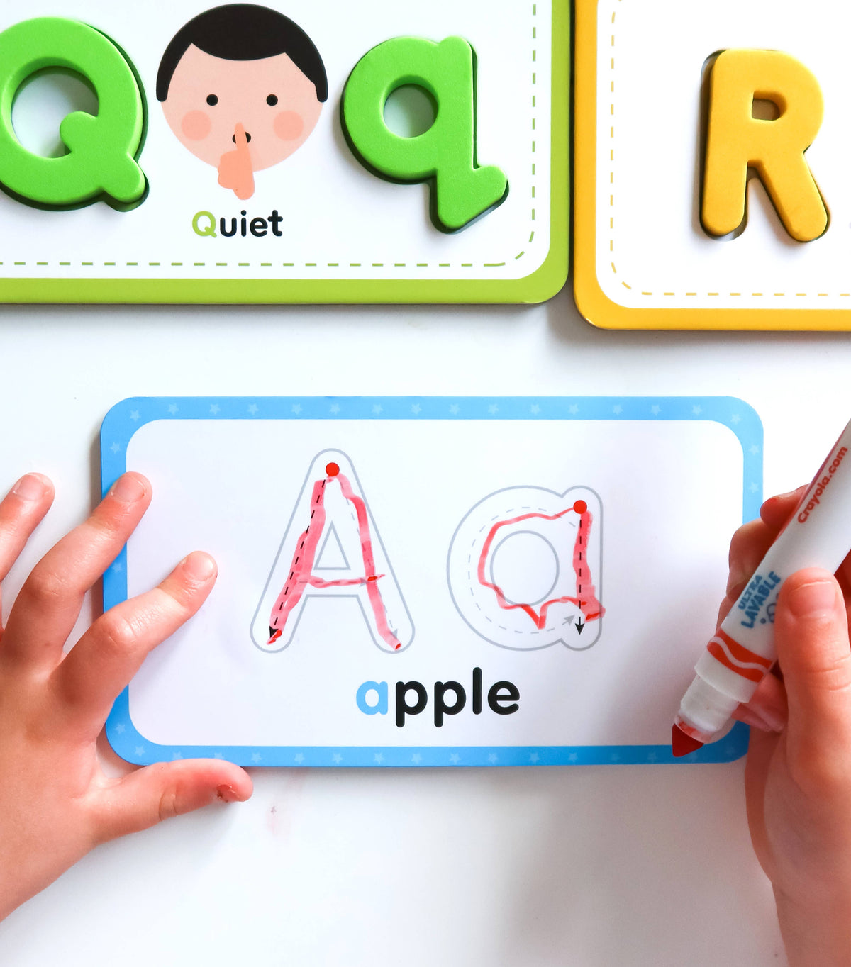 Children's educational board with letters Q, R, and a child's hand holding a marker near a card with 'A' and 'apple'.