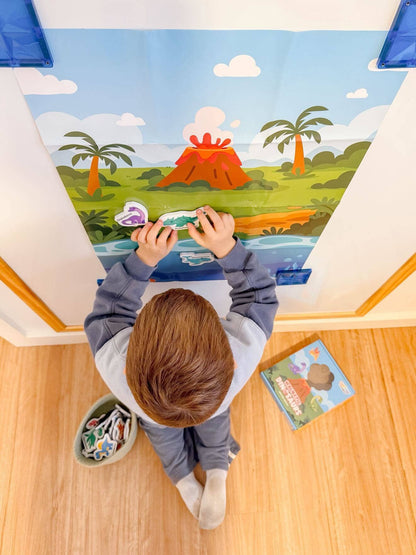A child viewed from above is placing colorful dinosaur stickers on a large illustrated wall mural of a prehistoric landscape with palm trees, water, and an orange volcano. A small book and a bin of stickers sit on the wooden floor nearby.