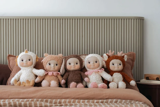 Five plush dolls dressed in animal costumes sit on a bed with brown pillows and a ribbed headboard in a softly styled bedroom.