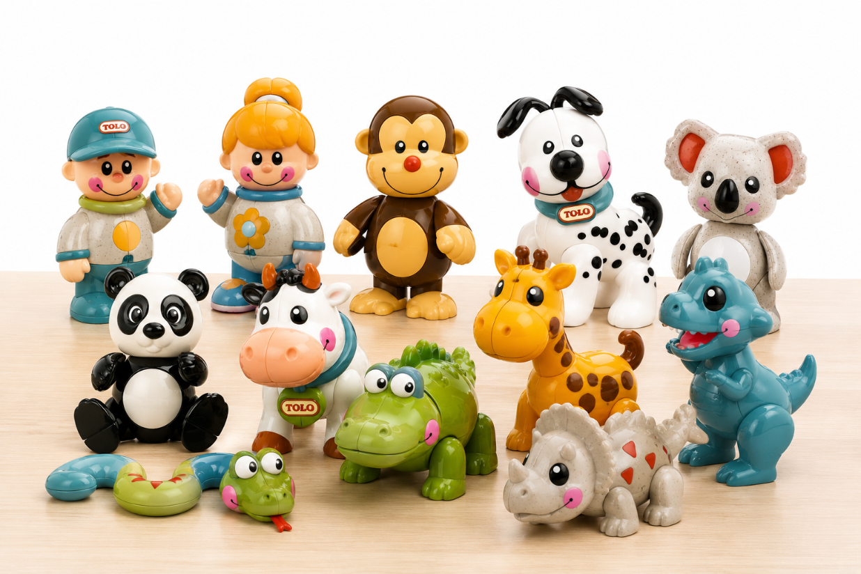 A set of colorful TOLO brand plastic animal and character toys arranged on a light wood surface against a white background. The collection includes a boy and girl astronaut figure, a monkey, a Dalmatian, a koala, a panda, a cow, a giraffe, a blue dinosaur, a green alligator, a triceratops, and a small snake.
