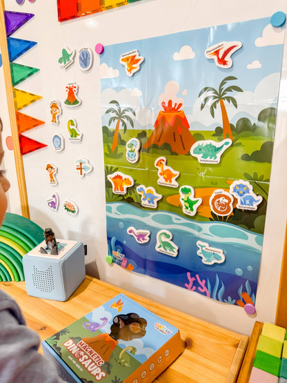 A colorful children's educational wall poster and matching dinosaur stickers displayed on a white wall in a classroom or playroom setting. A child's hand is visible at the bottom left, interacting with the toy.